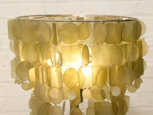 Load image into Gallery viewer, A modern chandelier or table lamp featuring a tiered design made of overlapping translucent shell-like discs in a warm gold and beige tone, illuminated from within against a white brick wall background.
