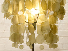 Load image into Gallery viewer, A hanging shell chandelier made of layered, translucent olive-green and beige seashells, illuminated from within against a white brick wall background.
