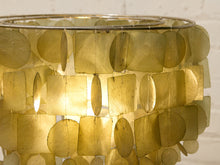 Load image into Gallery viewer, A close-up of a decorative lampshade made of overlapping translucent oval and rectangular leaf-like pieces, glowing warm yellow from an internal light source against a white wall background.
