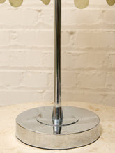 Load image into Gallery viewer, A close-up of a polished silver metal lamp base on a light beige stone surface, with a cream brick wall background.
