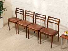 Load image into Gallery viewer, Four mid-century modern wooden dining chairs with brown leather seats, arranged in a row against a white brick wall on a beige carpet.
