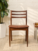 Load image into Gallery viewer, A mid-century modern wooden dining chair with a brown leather seat, positioned in front of a white brick wall with a potted plant on the left and a side table on the right.
