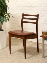 Load image into Gallery viewer, A mid-century modern wooden dining chair with a brown leather seat cushion, positioned next to a small side table on a textured beige rug against a white brick wall.

