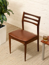 Load image into Gallery viewer, A mid-century modern wooden dining chair with a brown leather seat cushion, positioned on a beige textured rug next to a white brick wall, a potted plant, and a small side table.
