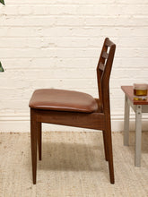 Load image into Gallery viewer, A mid-century modern wooden dining chair with a brown leather seat cushion, positioned next to a small side table against a white brick wall.

