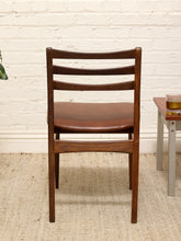 Load image into Gallery viewer, A mid-century modern wooden dining chair with a brown leather seat, positioned in front of a white brick wall on a woven jute rug.

