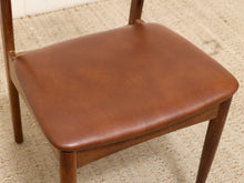 Load image into Gallery viewer, A close-up of a mid-century modern wooden dining chair with a warm brown leather upholstered seat cushion, set on a textured beige woven rug.
