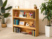Load image into Gallery viewer, A light natural wood 4-tier bookshelf styled with books, decorative figurines, a small clock, and a framed picture, set against a white brick wall with potted plants on either side.
