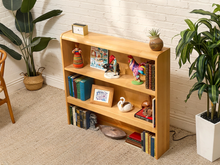 Load image into Gallery viewer, A light natural wood 4-tier bookcase styled with books, small figurines, and framed pictures, set in a room with a white brick wall, beige carpet, and potted plants.
