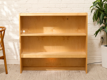Load image into Gallery viewer, A three-tier wooden bookshelf with a natural light oak finish, standing against a white brick wall with a potted plant and a chair partially visible.
