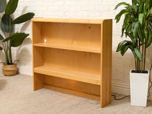 Load image into Gallery viewer, A light natural wood bookshelf with three shelves, positioned against a white brick wall on a beige carpet, flanked by potted green plants.
