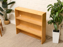Load image into Gallery viewer, A light natural wood three-tier bookcase standing on a beige carpet against a white brick wall, flanked by potted green plants.
