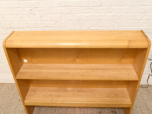 Load image into Gallery viewer, A light natural wood three-tier bookcase with a curved top edge, shown against a white painted brick wall and beige carpet.
