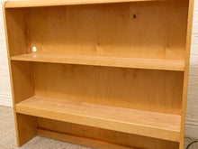 Load image into Gallery viewer, A light natural wood bookshelf with three shelves, photographed indoors against a white brick wall and carpeted floor.
