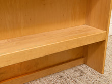 Load image into Gallery viewer, Close-up of a light natural wood bookshelf or shelf unit with a slatted front and open side compartments, shown against a beige carpeted floor.
