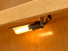Load image into Gallery viewer, A close-up of a warm white under-cabinet LED light fixture mounted on a light wood surface, casting a bright glow downward.
