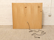 Load image into Gallery viewer, The back of a light wood cabinet or panel with a black power cord, standing against a white brick wall on a beige carpet.
