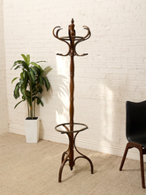 Load image into Gallery viewer, A tall dark brown wooden coat rack with multiple curved hooks, a central pedestal, and a lower ring stand, photographed in a room with a white brick wall, beige carpet, a potted plant, and part of a black chair visible.
