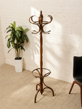 Load image into Gallery viewer, A tall dark brown wooden coat rack with multiple curved hooks, a central pedestal, and a circular lower shelf, shown in a room corner with a white brick wall, beige carpet, a potted plant, and part of a black chair.
