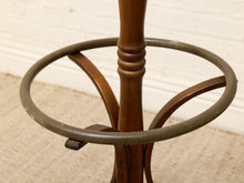 Load image into Gallery viewer, A close-up of a vintage wooden coat rack stand with a dark metal ring and curved wooden supports, set against a white brick wall and beige carpet.
