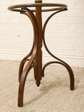 Load image into Gallery viewer, A vintage wooden pedestal stand with curved bentwood legs and a circular metal top ring, shown on a textured beige rug against a white brick wall.

