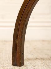 Load image into Gallery viewer, A close-up of a curved dark brown wooden leg or frame piece standing on a textured beige woven rug against a light cream wall.
