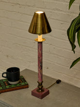 Load image into Gallery viewer, A table lamp with a warm brass-colored shade and base, a pink marbled central column, and a lit bulb, shown on a tabletop against a white brick wall with a plant and a black mug nearby.
