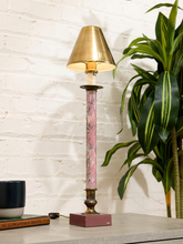 Load image into Gallery viewer, A decorative table lamp with a gold-toned metal shade and base, a pink floral-patterned central column, and a dusty rose rectangular base, shown lit on a tabletop next to a potted plant against a white brick wall.
