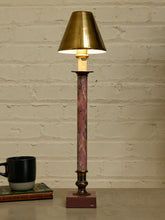 Load image into Gallery viewer, A vintage-style table lamp with a tapered brass shade, a pink and gray marbled column, and a dark metal base, shown lit on a tabletop against a white brick wall.
