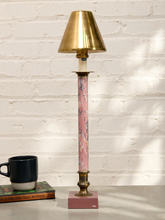 Load image into Gallery viewer, A tall table lamp with a polished brass shade and base, a pink marble-patterned column, and a pink square base, styled on a tabletop next to a black mug against a white brick wall.

