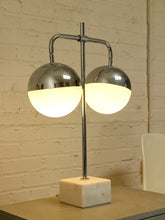Load image into Gallery viewer, A modern two-light table lamp with polished chrome metal arms and spherical shades, one of which is frosted white and glowing warm, mounted on a square white marble base against a cream brick wall.
