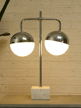 Load image into Gallery viewer, A modern two-light table lamp with polished chrome curved arms, glowing white globe shades, and a square white marble base, set against a beige brick wall.
