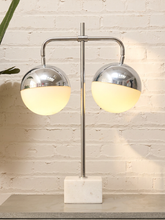 Load image into Gallery viewer, A modern two-light table lamp with polished chrome spherical shades and glowing warm white globe shades, mounted on a white marble base against a white brick wall.
