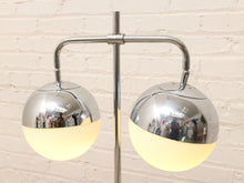 Load image into Gallery viewer, A modern two-light floor lamp with polished chrome metal arms and spherical shades, featuring a frosted warm white lower section on each globe, set against a light brick wall.
