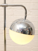 Load image into Gallery viewer, A modern floor lamp with a polished chrome spherical shade that is half reflective and half glowing warm white, mounted on a chrome stand against a light brick wall.
