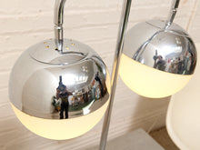 Load image into Gallery viewer, A modern two-light floor lamp with polished chrome spherical shades and warm white globe bulbs, shown lit against a light brick wall. The reflective surfaces show a photographer's reflection.
