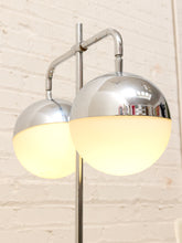 Load image into Gallery viewer, A modern two-light floor lamp with polished chrome metal arms and spherical shades, one of which is glowing warm white, set against a white painted brick wall.
