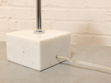 Load image into Gallery viewer, A close-up of a modern table lamp featuring a square white marble base with a visible power cord exit and a polished silver metal pole rising upward, set against a light brick wall background.
