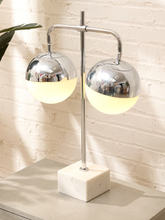 Load image into Gallery viewer, A modern two-light table lamp with polished chrome spherical shades, one glowing warm yellow, set on a white marble base against a white brick wall.
