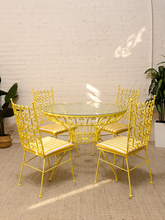 Load image into Gallery viewer, A 5-piece outdoor dining set featuring a round glass-top table and four matching yellow metal chairs with striped cushions, set against a white brick wall.

