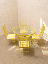 Load image into Gallery viewer, A yellow metal dining set consisting of a round glass-top table and four matching chairs with striped yellow and white cushions, set in a bright room with white brick walls and a beige carpet.
