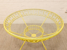 Load image into Gallery viewer, A bright yellow metal patio dining table with a decorative scrollwork frame and a clear glass top, set against a white brick wall and beige woven rug.
