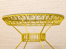 Load image into Gallery viewer, A bright yellow metal round table with an ornate scrollwork design, a glass top, and a lower shelf, set against a white brick wall background.
