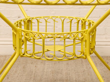 Load image into Gallery viewer, Close-up of a bright yellow metal garden table frame with a circular wire basket shelf and decorative scrollwork, set against a white brick wall and beige floor.
