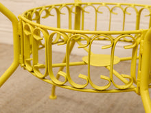 Load image into Gallery viewer, A close-up of a bright yellow metal garden furniture piece, showing decorative scrollwork and legs on a beige carpeted floor.
