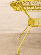 Load image into Gallery viewer, A bright yellow metal side table with an ornate scrollwork design, a glass top, and three legs, shown against a white brick wall and beige rug.
