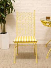 Load image into Gallery viewer, A yellow metal dining chair with a striped yellow and white cushioned seat, set against a white brick wall with a potted plant and a matching yellow table partially visible.
