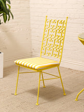 Load image into Gallery viewer, A yellow metal dining chair with an ornate scrollwork backrest and a yellow-and-white striped cushioned seat, shown in a bright room with a white brick wall and beige carpet.
