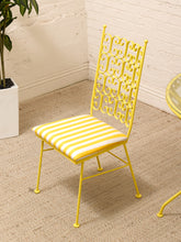 Load image into Gallery viewer, A yellow metal dining chair with an ornate scrollwork backrest and a yellow-and-white striped cushioned seat, shown in a room corner on a beige rug next to a white brick wall and potted plant.
