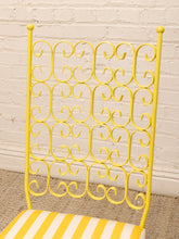 Load image into Gallery viewer, A close-up of a bright yellow metal chair frame with an ornate scrollwork backrest and a yellow-and-white striped cushion, set against a white painted brick wall and woven rug.
